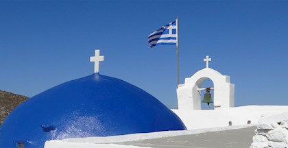 Toppen af en græsk kirke med det ikoniske blå kuppeltag med et kors på toppen, samt det græske flag og en kirkeklokke